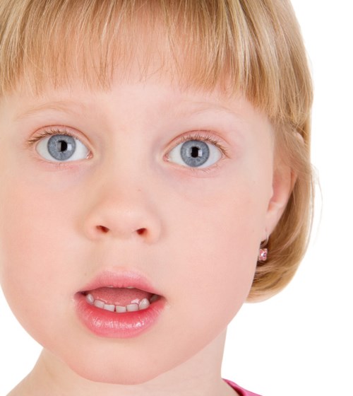 A young girl with blue eyes and blonde hair looks surprised or curious. She is wearing earrings and has slightly open lips.