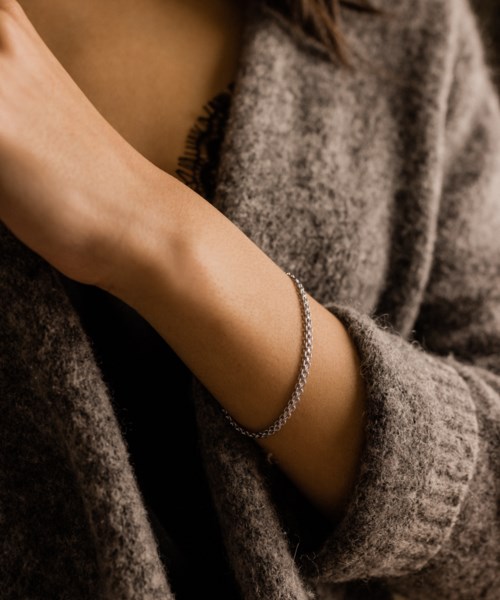 A person wears a silver bracelet and a cozy grey sweater. The focus is on the arm and accessories.
