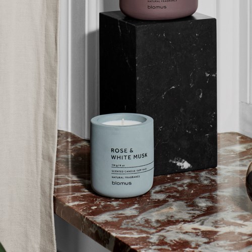 Blue candle labeled 'Rose & White Musk' on a marble surface, with a black pedestal and pink container in the background.