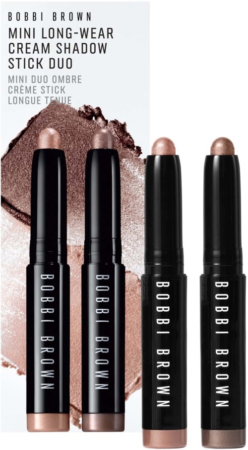 Vier Bobbi Brown oogschaduwstiften met zwarte handvatten en beige uiteinden, geplaatst voor bruine en beige oogschaduwmonsters.