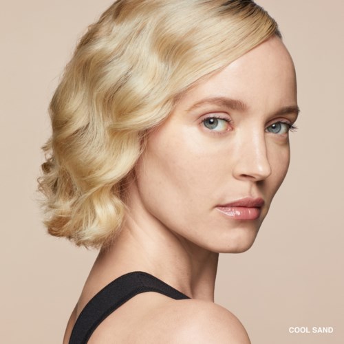 Eine Frau mit Lockenblondem Haar und blauen Augen blickt über ihre Schulter, trägt ein schwarzes Top und dezentes Make-up.