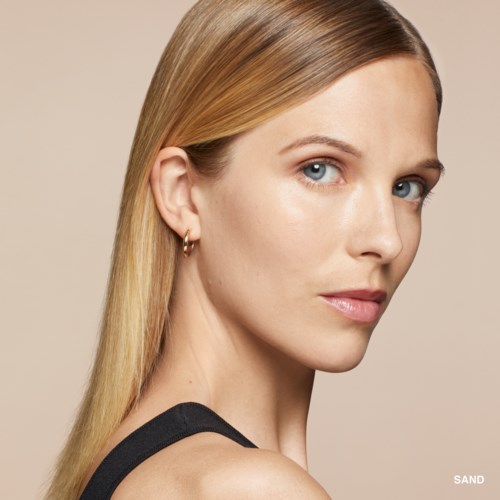Eine Frau mit langem blondem Haar, heller Haut, blauen Augen, kleinen Ohrringen und natürlichem Make-up schaut über die Schulter.