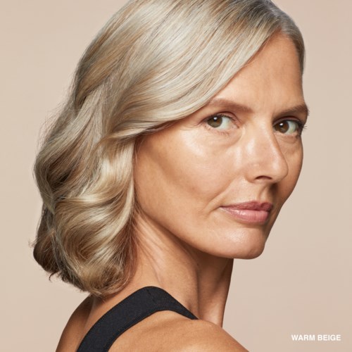 Eine Frau mit blonden, lockigen Haaren schaut über die Schulter vor einem beigefarbenen Hintergrund.