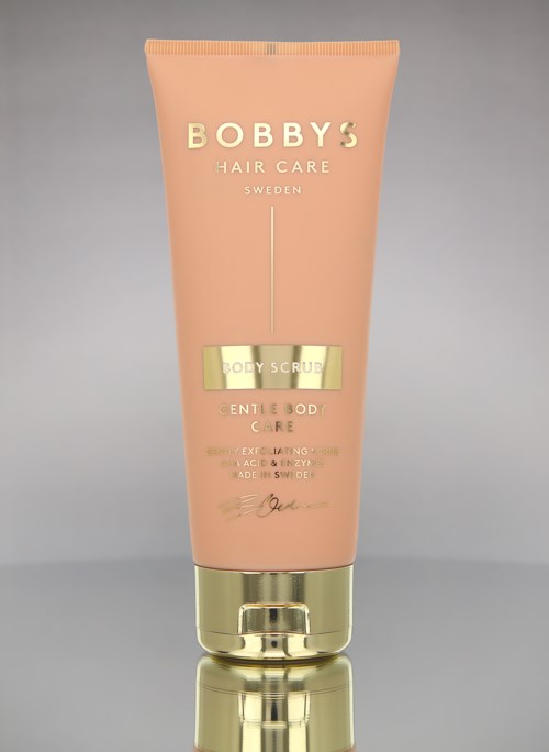 Pink tube med Bobby's Hair Care kroppsskrubb med gullfarget lokk og tekst, plassert på en grå bakgrunn.