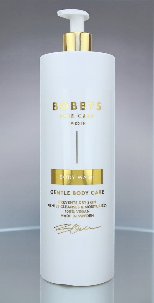 En hvit flaske med gullfargede bokstaver, pumpehode, merket 'BOBBYS Hair Care' og 'Body Wash', på en grå bakgrunn.