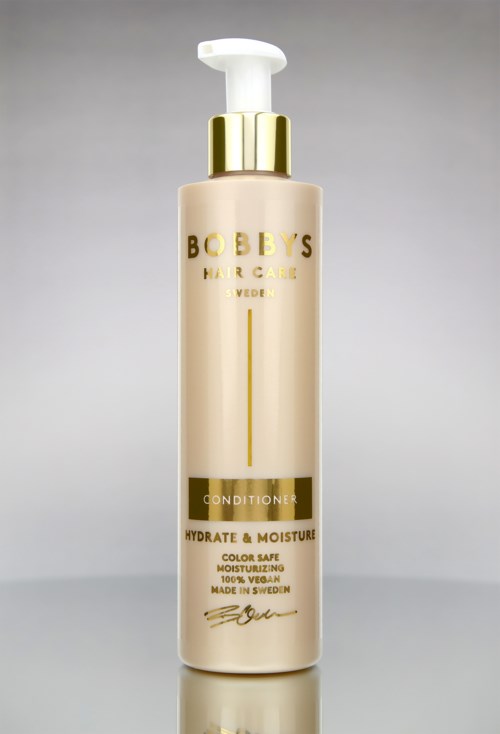En flaska Bobby's Hair & Care Conditioner med gul och vit design, som visar att den ger fukt och återfuktar håret.