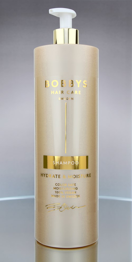 En lång, beige schampoflaska med vit pump, guld accenter och svart och guld text.
