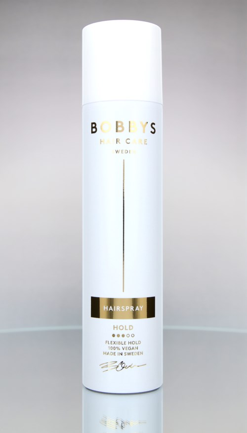 En lång, vit sprayflaska med gyllene och svarta text, märkt "Bobby's Hairspray" och "HOLD".