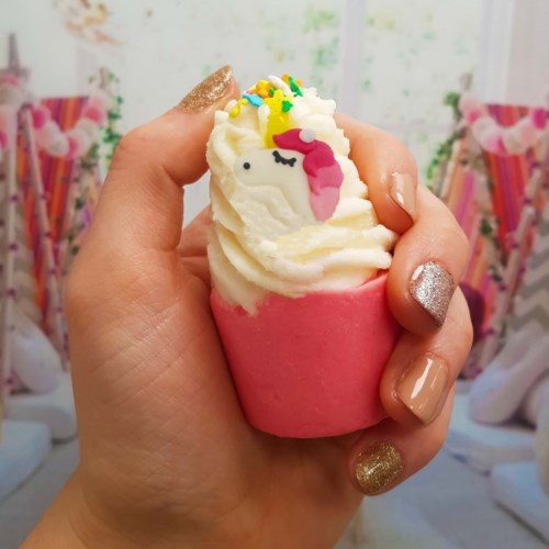 En hånd holder en rosa cupcake med hvit frosting, en enhjørningdekorasjon og fargede strøssel.