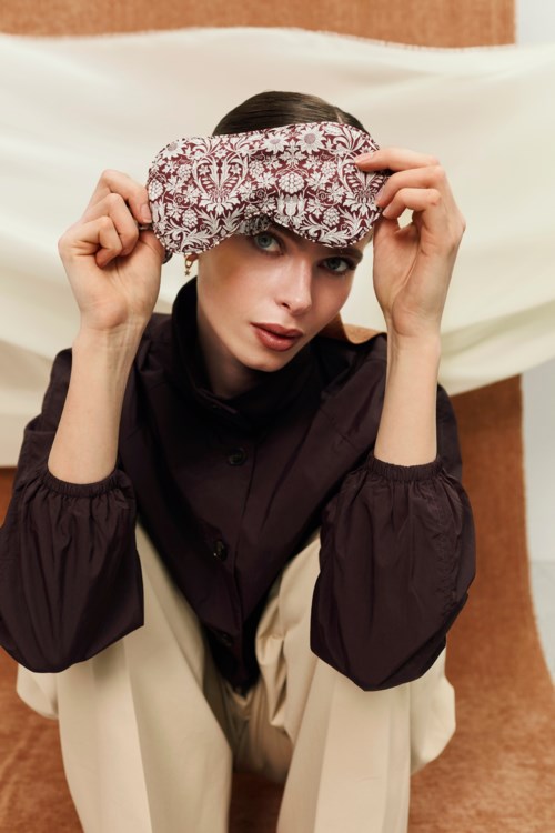 En kvinna poserar med en paisley-sovmask på huvudet, bär en mörk blus och beige byxor, mot en enkel bakgrund.