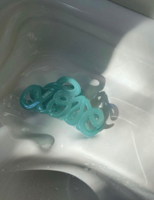 Ett turkost plastföremål med böjda ändar ligger i en vit handfat, med skuggor från ljuset.