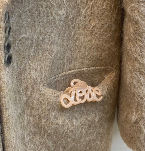 A beige coat with a pocket holding a 'love' pin or badge.