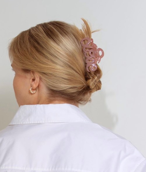 Frau mit blonden Haaren in Hochsteckfrisur, befestigt mit rosa Haaraccessoire, trägt Ohrringe und ein weißes Hemd.
