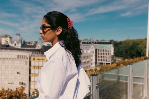 Eine Frau mit dunklem Haar, Sonnenbrille und einem pinken Haaraccessoire steht draußen an einem sonnigen Tag und schaut auf die Stadt.