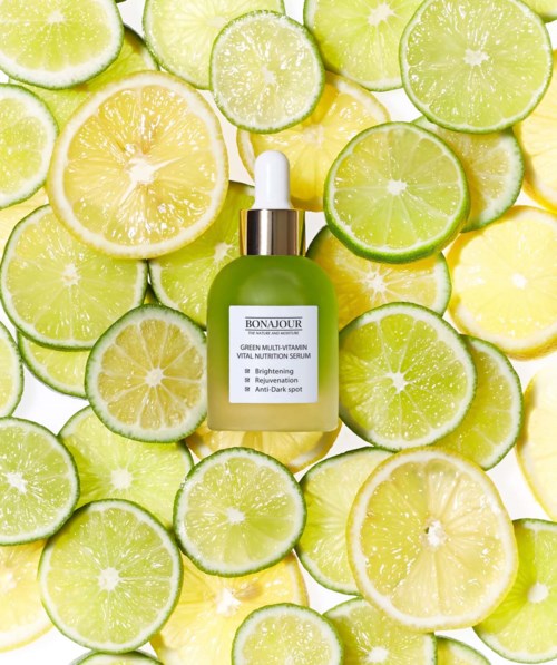 A green serum bottle surrounded by lemon and lime slices. The label mentions vitamins and brightening effects.