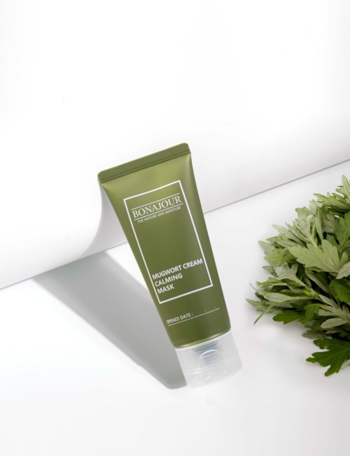 En grønn tube med Bonjour Mugwort Cream Calming Mask og en grønn plante ved siden av på hvit bakgrunn.