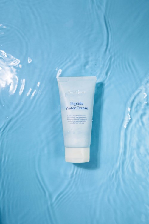 A white tube of peptide water cream floats on clear blue water with ripples. The background is light blue.