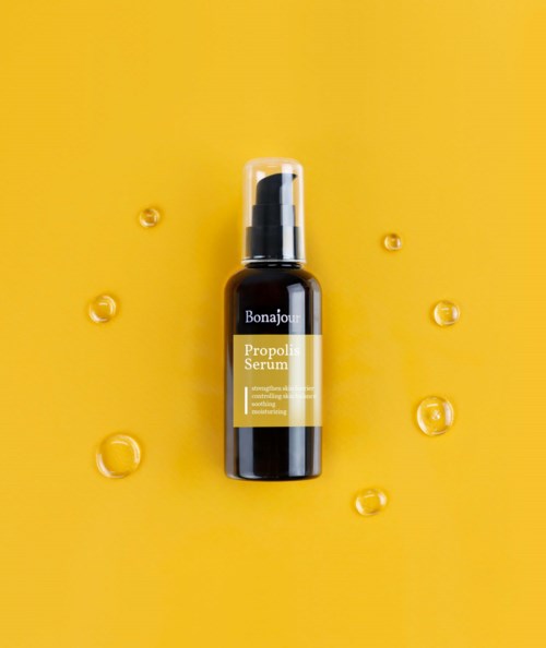 Een zwarte fles met label 'Propoli Serum' door Bonajour op een gele achtergrond, met kleine gele druppels eromheen.
