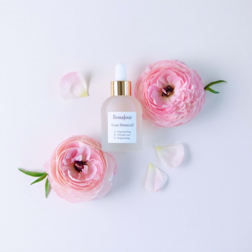 Een huidverzorgingsfles omgeven door roze bloemen en verspreide bloembladeren op een witte achtergrond.