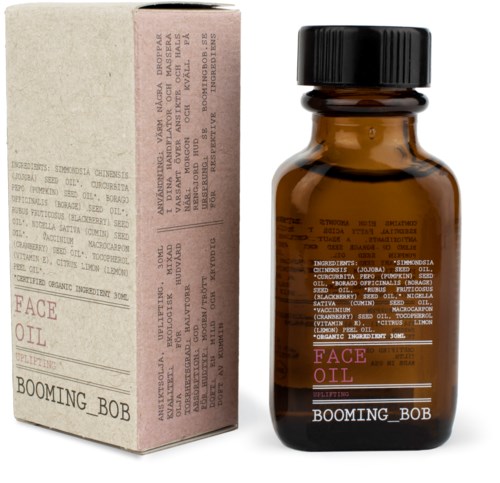 Brązowa butelka z etykietą 'Face Oil' i obok niej różowa i beżowa pudełko.
