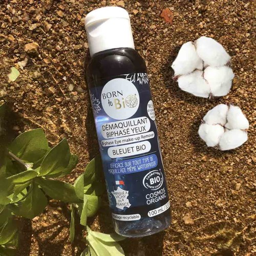 Black bottle of eye makeup remover on dirt, with cotton and green leaves nearby.