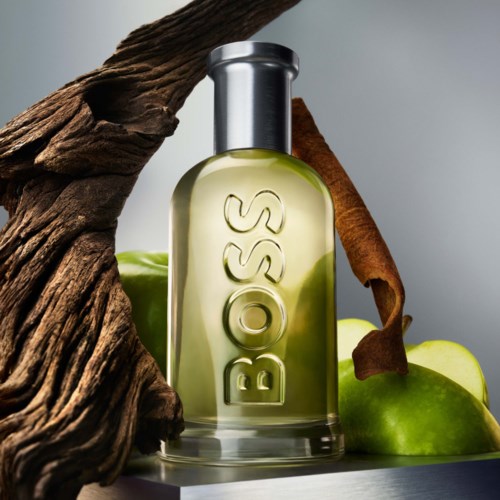 A clear perfume bottle with the brand name 'BOSS' surrounded by apples, wood, and cinnamon sticks.