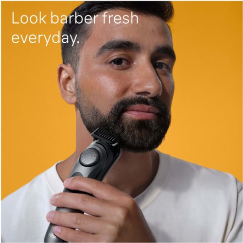Een man met een baard scheert zijn baard met een elektrische scheerapparaat tegen een gele achtergrond.