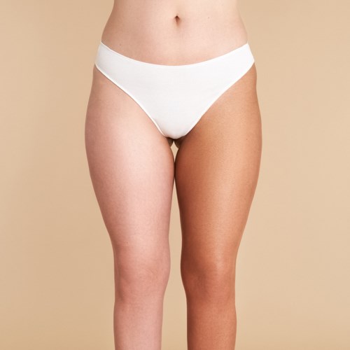 En person med ett ben mørkere enn det andre, iført hvitt undertøy, står mot en beige bakgrunn.
