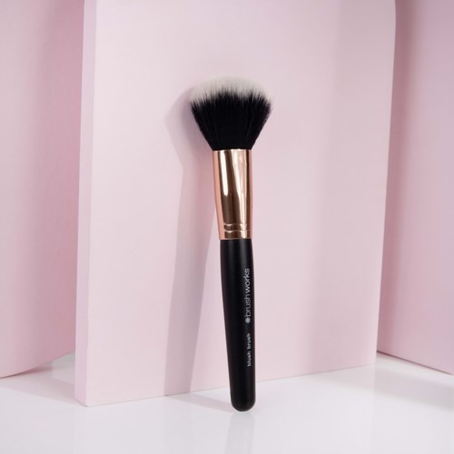 En makeupborste med svart handtag, guldfäste och fluffiga vita och svarta borststrån, luta mot rosa bakgrund.