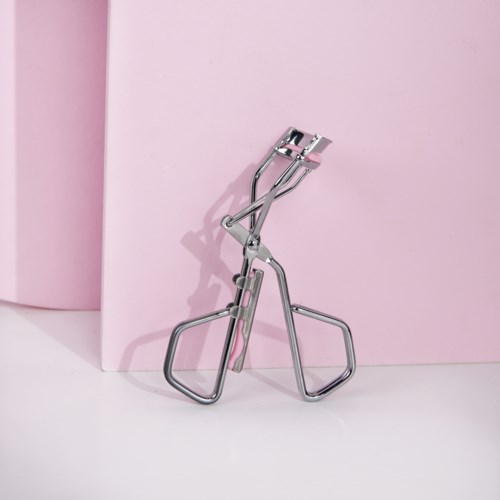 Een metalen wimperkruller staat voor een roze achtergrond, met schaduw op de muur.