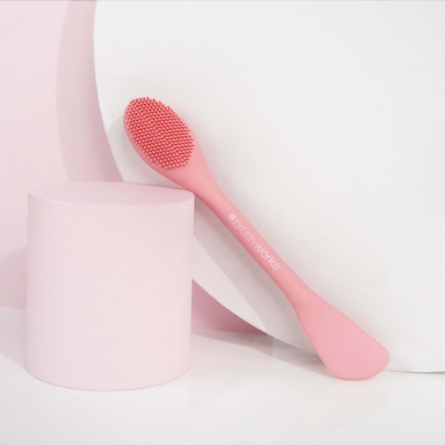 En rosa silikon ansiktsbørste hviler mot en hvit og rosa minimalistisk bakgrunn.