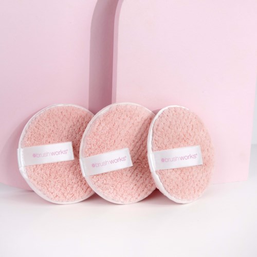 Drie roze reinigingspads met witte banden met de tekst "#brush works" liggen voor een roze achtergrond.