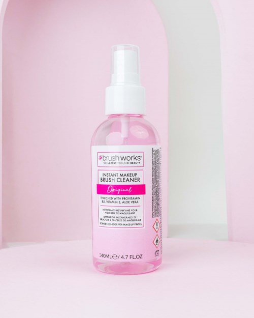 En pink flaske med makeup børsterenser med spray, placeret mod pink og hvid baggrund.