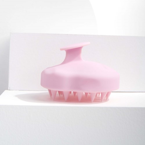 A pink scalp massager with soft rubber bristles and a handle on top, placed on a white surface.