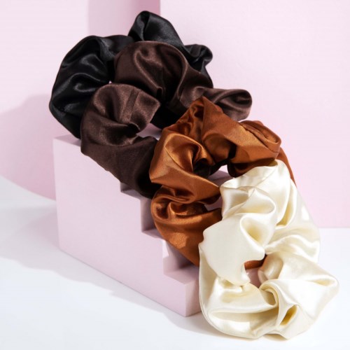A display of four satin scrunchies in black, brown, rust, and cream, arranged on a pink block against a pink background.
