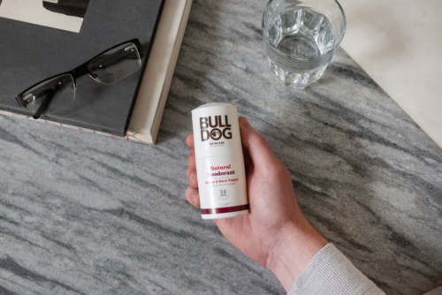 Hånd holder en deodorant stick mærket 'BULLDOG' på et bord med briller og bøger.