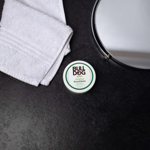 Ein weißes Handtuch, ein runder Spiegel und eine Dose Bartbalsam auf einer dunklen Oberfläche.