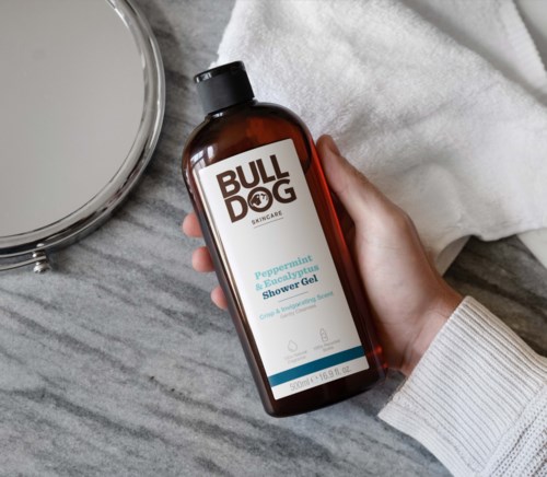 Käsi pitää ruskeaa pulloa, jossa lukee "BULLDOG" ja suihkugeeli, vieressä peili ja valkoinen pyyhe.