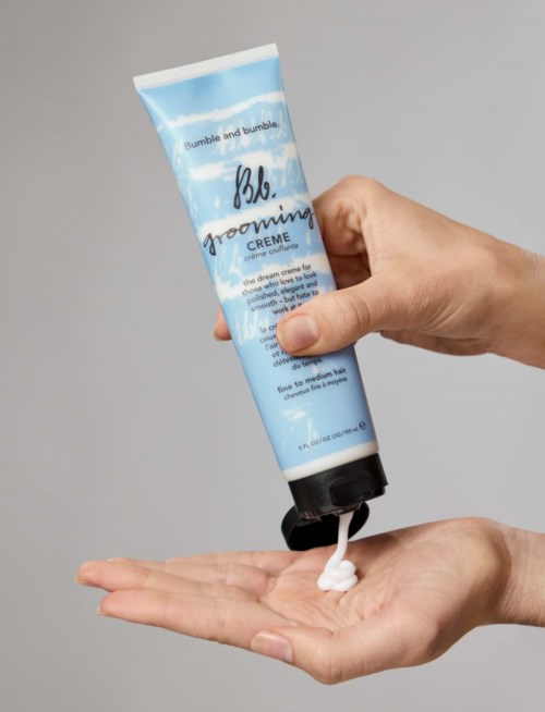 A hand holds a blue tube of Bumble and Bumble grooming cream, dispensing some white cream onto the hand.