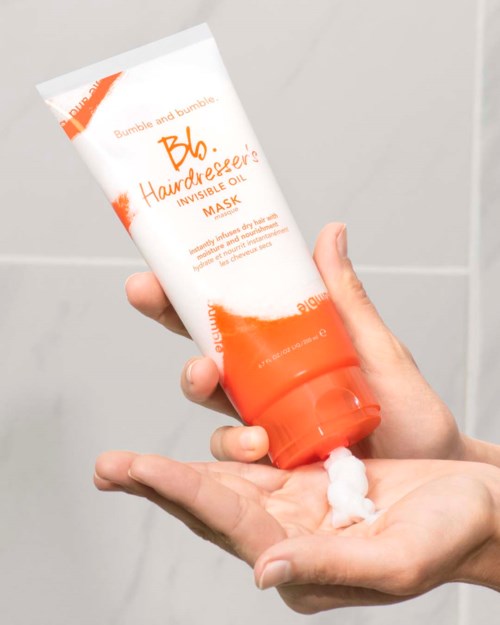 En person påfører en hvit krem fra et rør merket 'Bumble and bumble. Hairdresser's Invisible Oil Mask' på hånden.