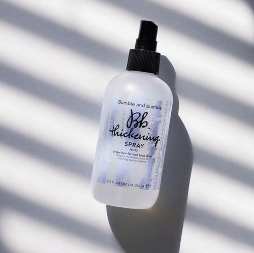 Biała butelka spray z oznaczeniem 'Bumble and Bumble' i 'Thickening Spray' z czarnym dyszelem, cień z liniami.
