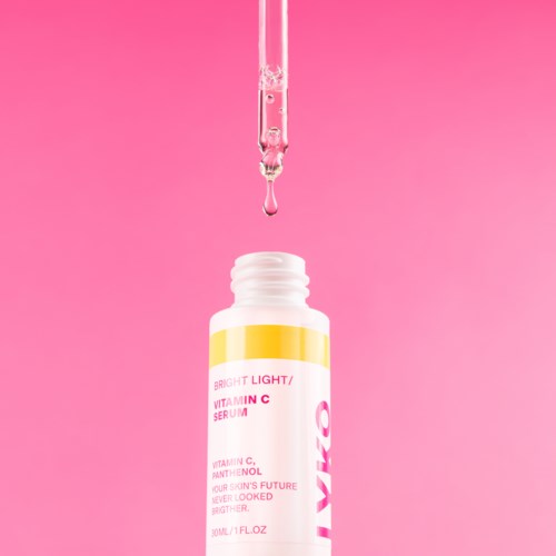A dropper releases serum into a small white bottle with a colorful label, set against a pink background.