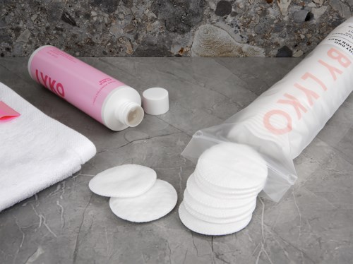 Bomullspads, en rosa flaska, en handduk och en plastpåse är på ett grått arbetsbord.