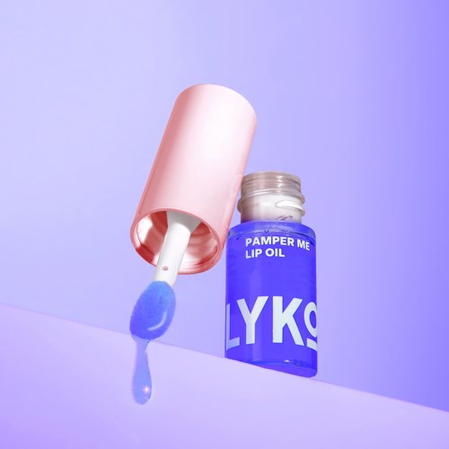 En blå flaska märkt 'Pamper Me Lip Oil' med applicator, ligger på lila bakgrund.