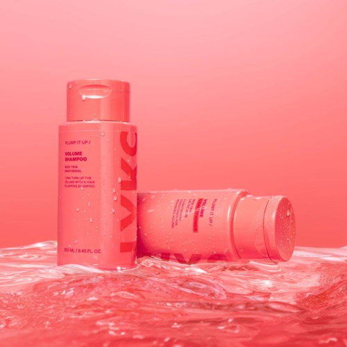 Two pink bottles of shampoo placed on a wet surface with water around them, bright background.