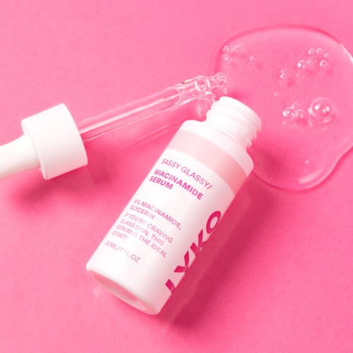 A skincare serum bottle with a dropper, lying on a pink surface with some serum spilled and bubbles.