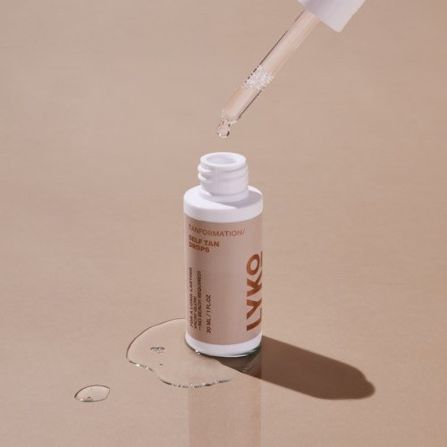 A white bottle labeled 'self tan drops' with a dropper releasing liquid, and some liquid spilled on beige surface.