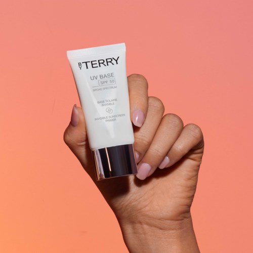 Hand som håller i en tub med UV-kräm med rosa bakgrund, etiketten visar SPF 50 och varumärket BY TERRY.