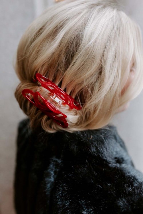 A child with blonde hair has a large red hair clip on the side of her head.