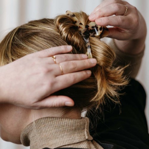 Eine Person fixiert die Haare einer Frau mit einer Haarspange, eine weitere Person assistiert. Ziel ist eine ordentliche Frisur.